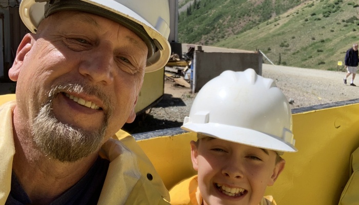 A smiling man and young boy wearing white hard hats and yellow jackets outdoors.