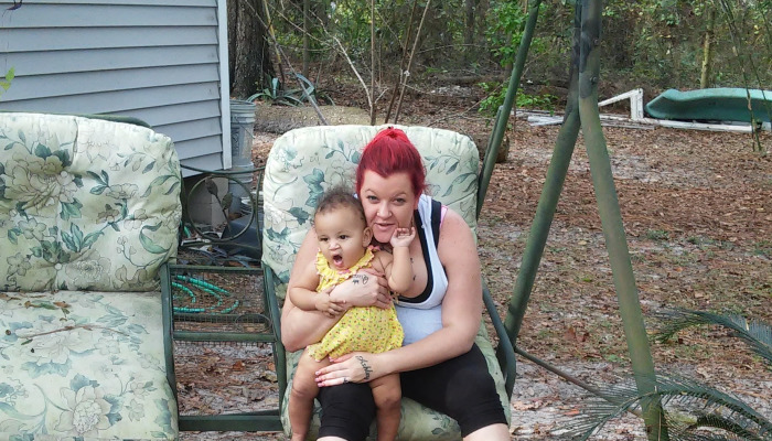 A woman with bright red hair holds a toddler in a yellow dress while sitting on a floral porch swing outdoors.