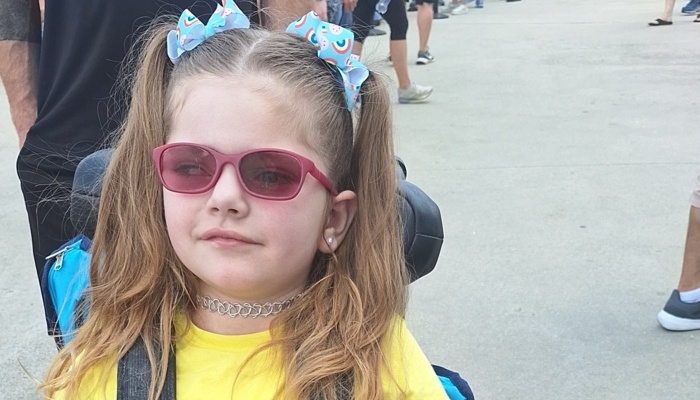 Young girl with pigtails, pink sunglasses, and a yellow shirt sitting outdoors in a wheelchair.