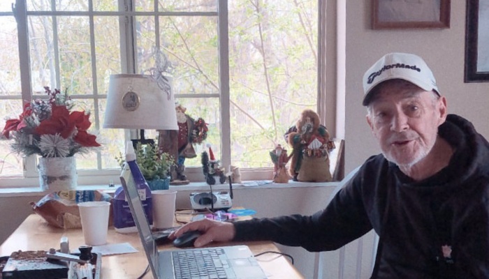 An elderly man wearing a white cap sits at a desk near a window, smiling while using a laptop.