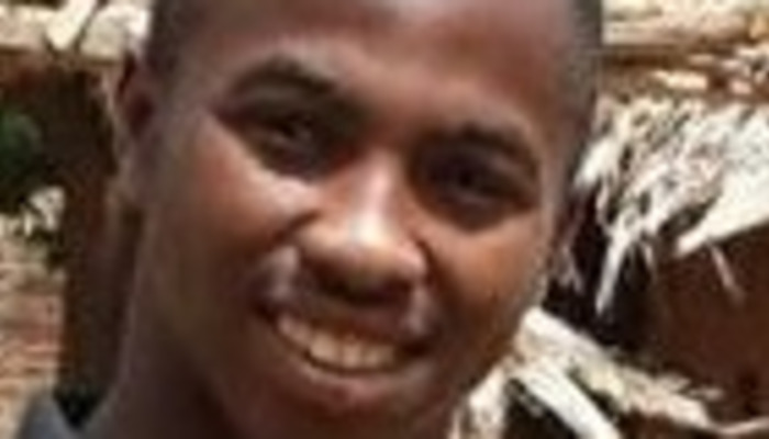 Close-up portrait of a young Black man smiling warmly in front of a rustic structure.
