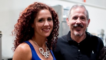 A smiling woman with curly red hair and a silver necklace next to a smiling man with a mustache.
