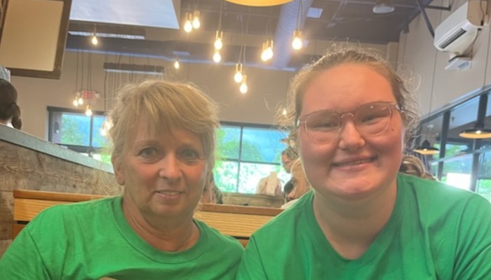 A blonde woman and a young woman with glasses smiling in matching green t-shirts inside a restaurant.