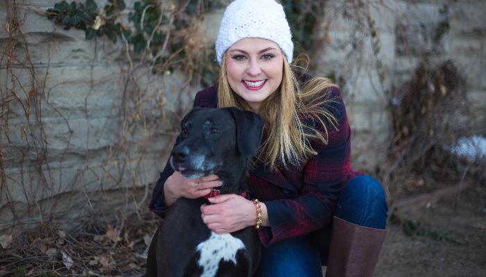 A smiling woman in a white beanie and plaid coat hugging her black dog outdoors against a stone wall.