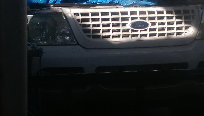 Front grille, logo, and headlight of a white Ford vehicle covered by a blue tarp.