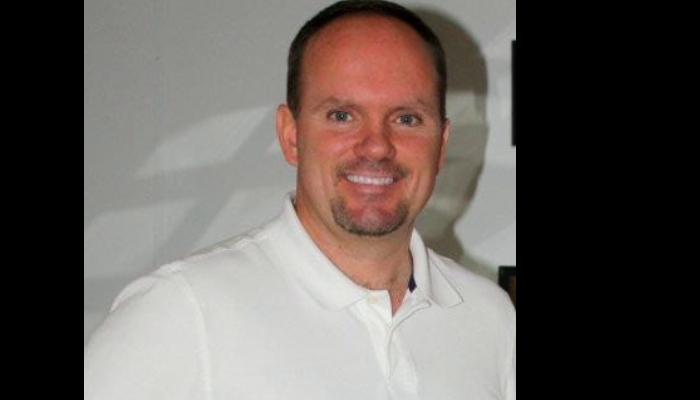 A smiling man with short hair and a goatee wearing a white polo shirt.