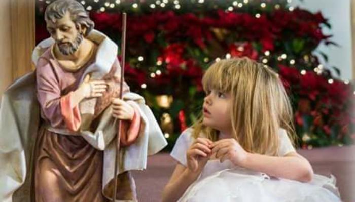 A young girl in a white dress looks up at a nativity statue of Saint Joseph in front of Christmas poinsettias.