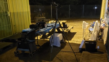 Telescope on a tripod set up at night beside a picnic table with gear, a pet stroller, and a cat nearby.