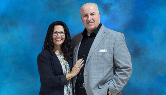 A smiling couple posing together against a blue backdrop, with the woman resting her hand on the man's grey blazer.