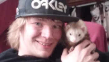 A smiling young man wearing an Oakley cap holds his pet ferret close to his face.