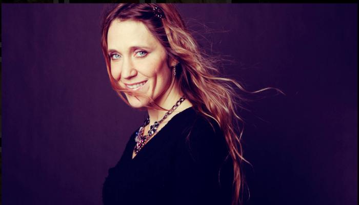 Smiling woman with blue eyes and long, wind-blown hair wearing a statement necklace.