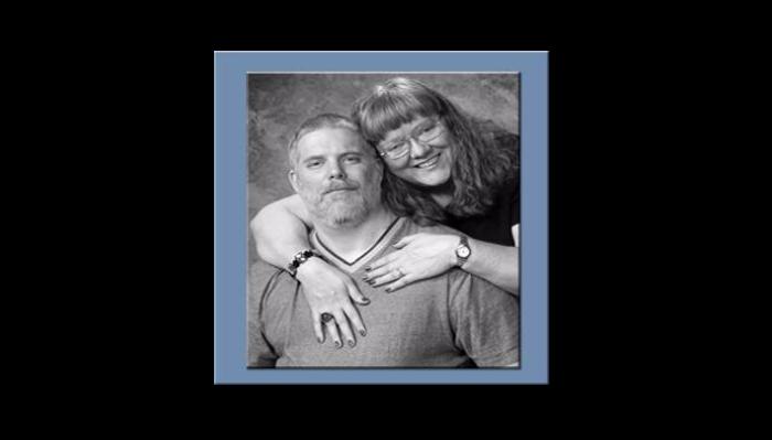Black-and-white portrait of a smiling woman embracing a bearded man from behind.