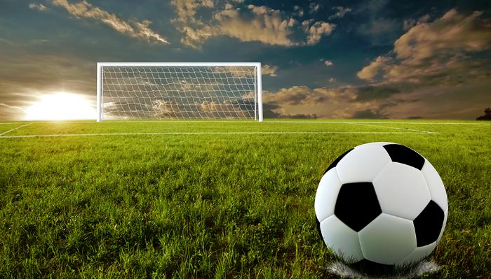 A soccer ball resting on a green pitch facing a goal net under a dramatic sunset sky.