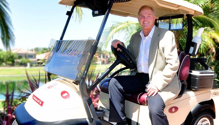 A smiling man in a suit jacket sitting in a golf cart on a sunny golf course.