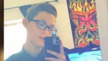 Young person with short hair and glasses taking a mirror selfie with a smartphone.