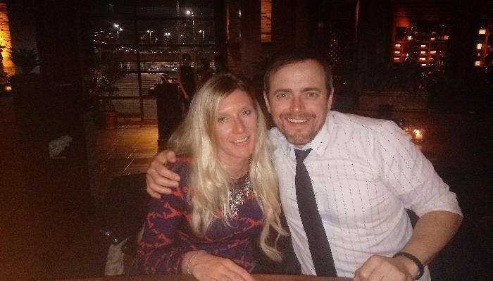 A smiling man with his arm around a blonde woman, posing together in a dimly lit restaurant at night.