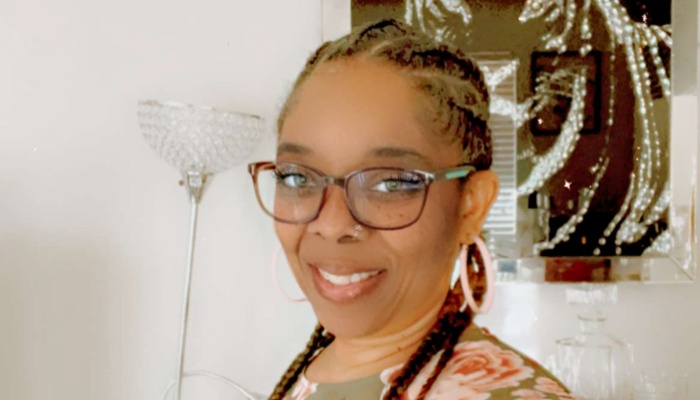 A smiling Black woman with braided hair, glasses, and pink hoop earrings.