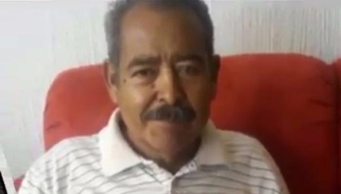 Middle-aged man with a dark mustache wearing a striped polo shirt, sitting in front of a red couch.