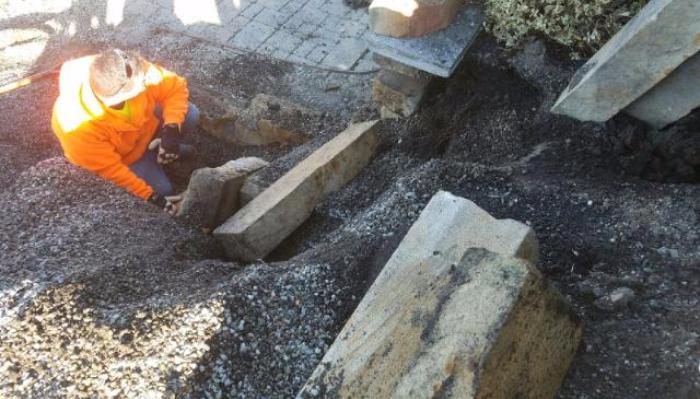 A worker in a bright orange jacket installs heavy stone blocks to construct outdoor steps.