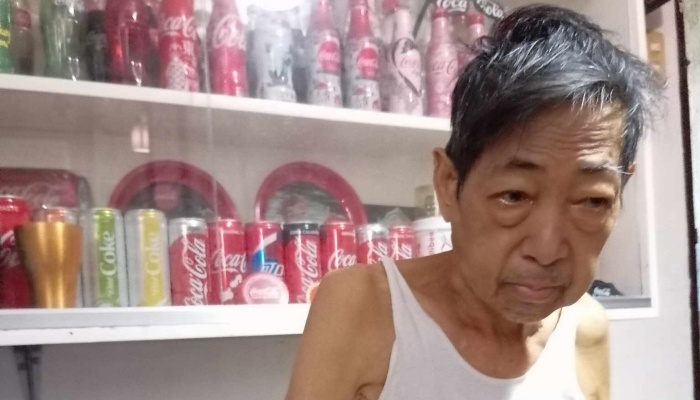 An elderly Asian man wearing a white tank top in front of shelves filled with Coca-Cola collectibles.