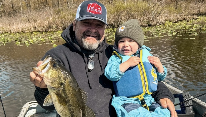 A smiling man holds a large bass on a boat alongside a young toddler wearing a blue life vest and green beanie.