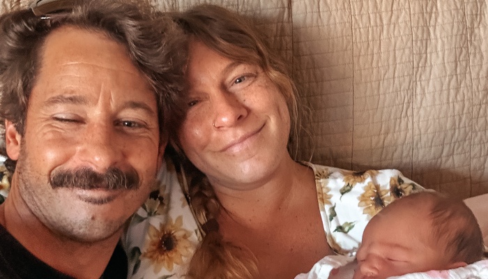Smiling parents pose closely together while holding their sleeping newborn baby.