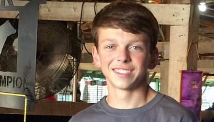 A smiling teenage boy with brown hair and blue eyes wearing a grey t-shirt in a barn.