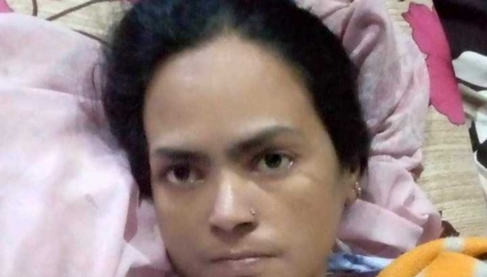 A woman with dark hair and a nose stud lies in bed, looking solemnly at the camera.