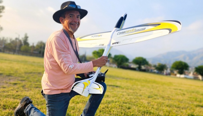 A smiling man wearing a hat kneels in a grassy field while holding a white and yellow RC glider airplane.