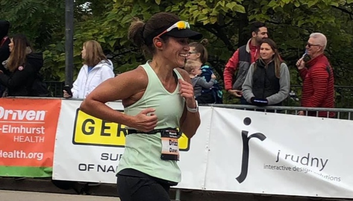 A smiling woman wearing a visor and race bib runs past cheering spectators during a marathon.