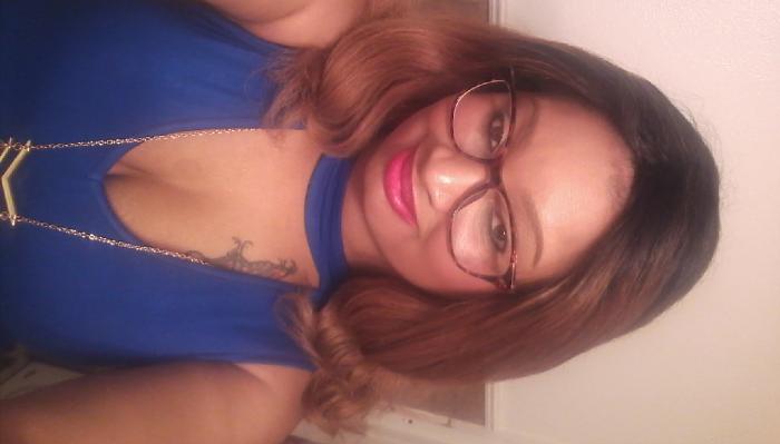 Smiling woman with wavy brown hair, glasses, and pink lipstick wearing a blue top and gold necklace.