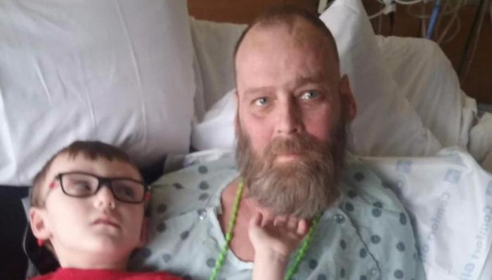 A bearded man wearing a hospital gown lies in bed next to a young boy with glasses.