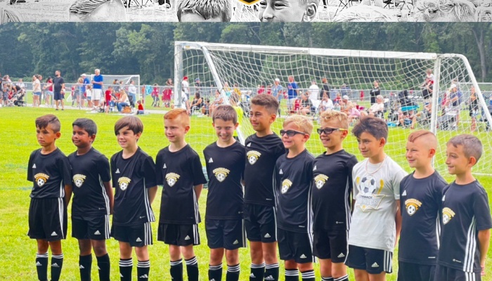 A row of young boys in black soccer uniforms standing on a grassy field in front of a soccer goal.