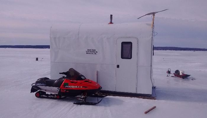 A red snowmobile parked next to a white ice fishing shack on a frozen, snowy lake.