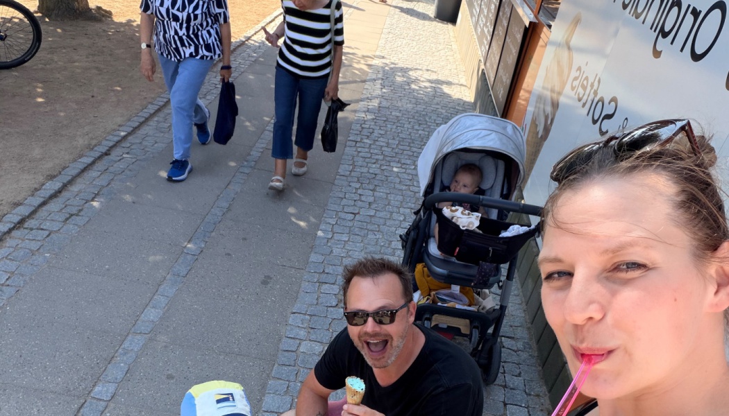 A woman takes a selfie outdoors beside a smiling man holding an ice cream cone and a baby in a stroller.