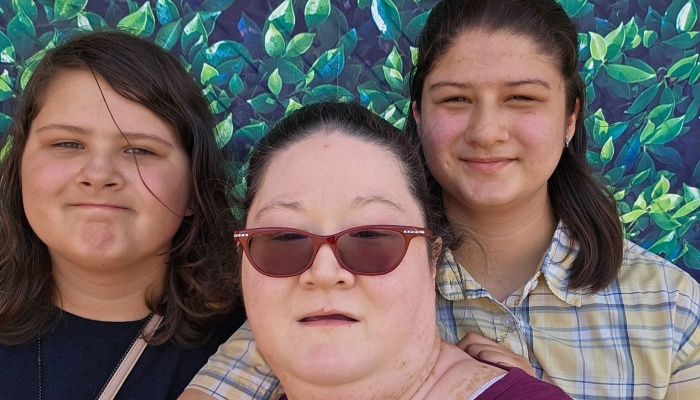 A woman wearing red glasses poses with two smiling girls in front of a leafy green background.