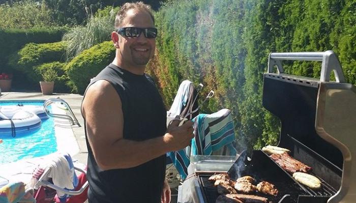 A smiling man in sunglasses grills meat on an outdoor barbecue next to a backyard swimming pool.