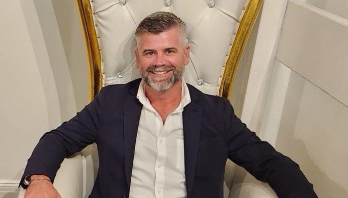 Smiling man with a grey beard in a dark suit sitting in an ornate white and gold throne chair.