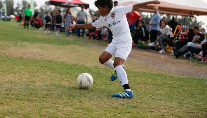 A young boy in a white uniform dribbles a soccer ball across a grass field during a match.