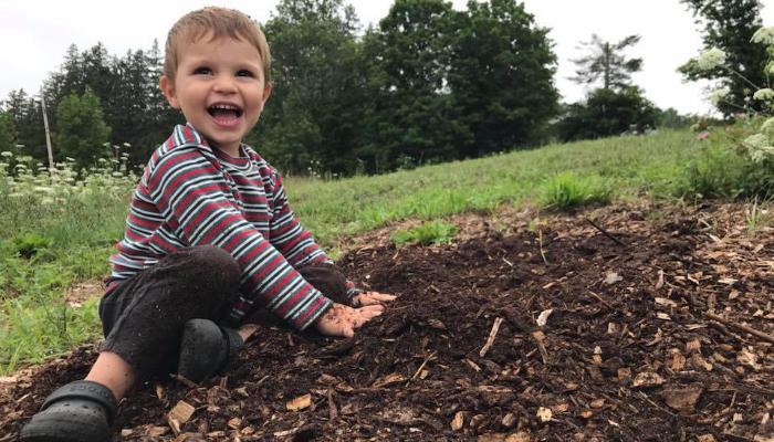 A joyful toddler in a striped shirt sits outdoors, laughing while playing in a pile of dark soil.
