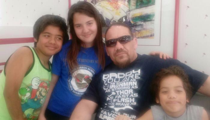 A father wearing sunglasses poses warmly with his three smiling children.