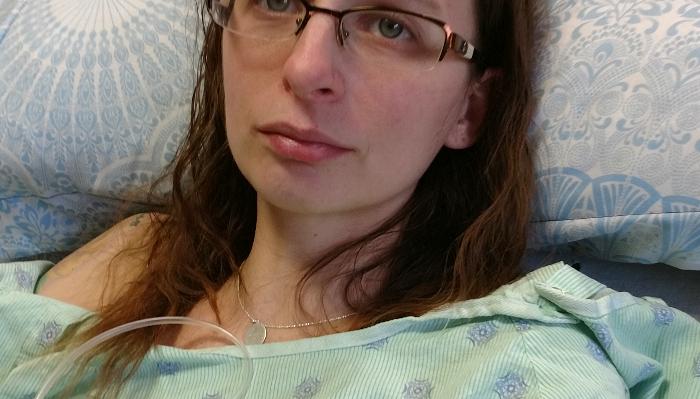 A woman with glasses wearing a green hospital gown lies in bed with medical tubing attached.