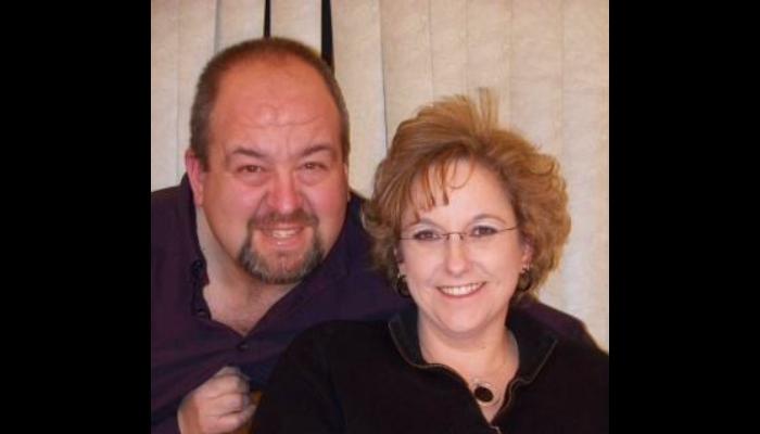 A smiling middle-aged couple posing close together, a man with a goatee beside a woman with glasses.