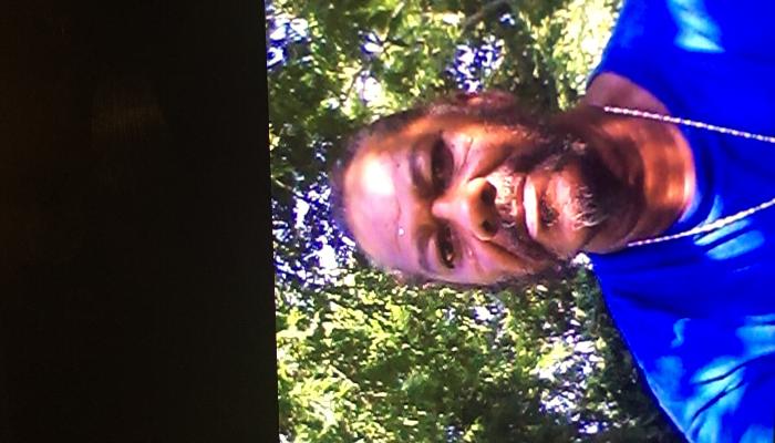 A man with a beard and silver chain wearing a blue shirt outdoors with green trees in the background.