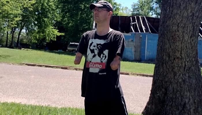 Man with amputated forearms wearing a cap and t-shirt, standing outdoors in front of a damaged building.