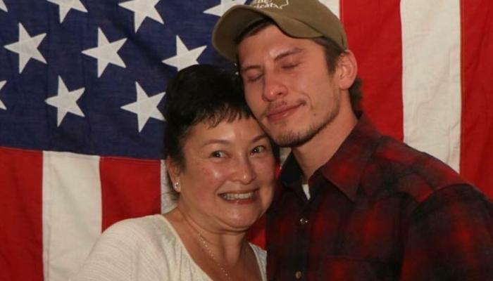 A smiling woman and a young man in a cap and plaid shirt pose together in front of an American flag.
