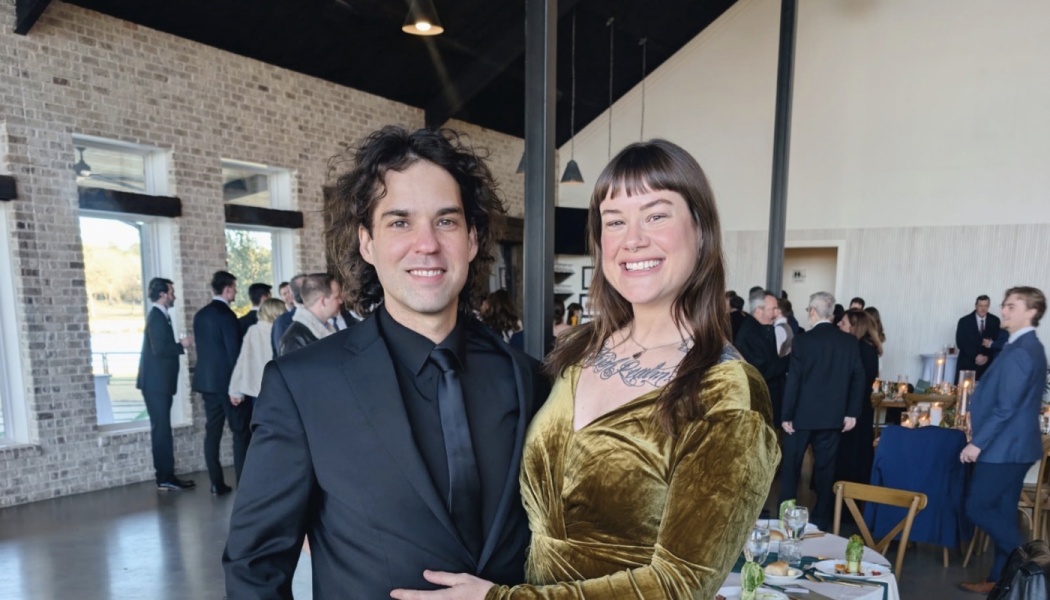 A smiling man in a black suit and a woman in an olive green velvet dress pose together at a formal indoor event.