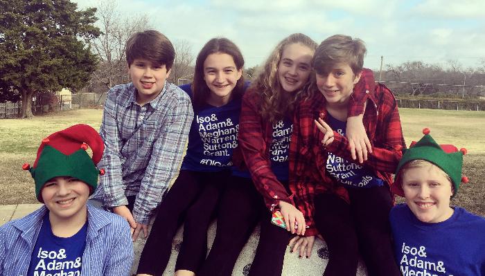 Six smiling kids sitting outside together, wearing matching blue t-shirts and two festive elf hats.
