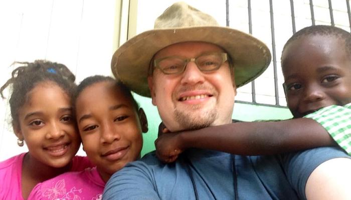 A smiling man in a wide-brimmed hat takes a close selfie surrounded by three happy young children.