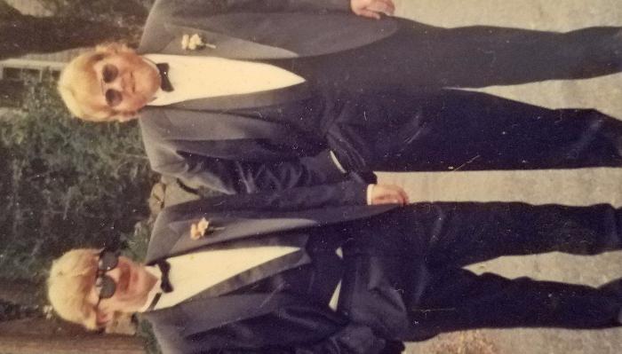 Two people wearing matching tuxedos, bow ties, and sunglasses standing side by side outdoors.